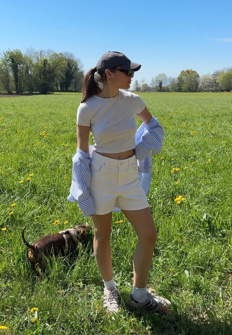 Femme en short blanc et crop top debout dans un champ herbeux avec des fleurs jaunes, portant une casquette et des lunettes de soleil, avec un petit chien brun à proximité.