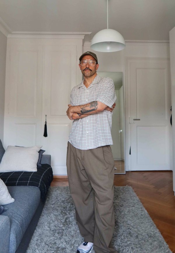 Mann mit Brille, Kappe, Tattoos und Schnurrbart steht mit verschränkten Armen in einem Wohnzimmer mit weißen Wänden, grauem Sofa und einer Hängeleuchte.