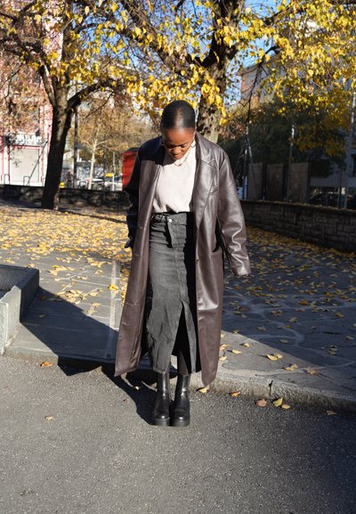 Abrigo largo de cuero marrón, suéter gris acanalado, falda de mezclilla negra con abertura frontal y botas de plataforma negras; hojas de otoño en el fondo.
