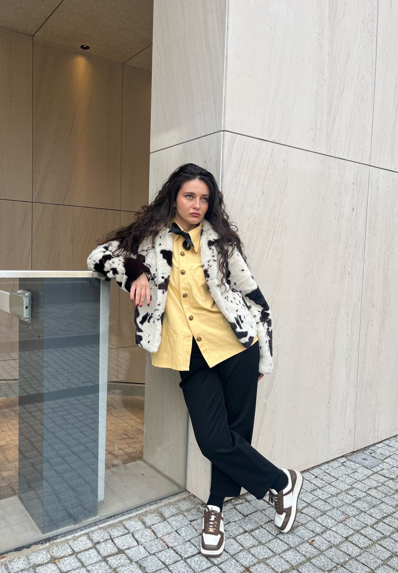 Viltotas kažokādas mētelis ar melniem un baltiem rakstiem, dzeltens krekls ar pogām, melnas svītrainas bikses un baltbrūni sporta apavi. Stāv pret sienu.