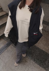 Juodas liemenė su Nike logotipu, šviesiai rudas megztinis su "EMESTUDIOS" tekstu, pilkos kelnės ir šviesūs batai. Juostelės tekstūra ant liemenės.