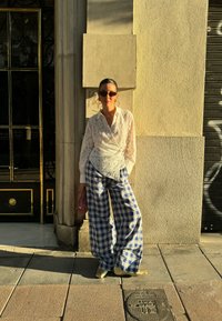 Λευκή δαντελένια μπλούζα με μακριά μανίκια, μπλε και λευκό καρό παντελόνι φαρδύ, διάφανα κίτρινα παπούτσια και ροζ τσάντα, μπροστά από έναν υφασμάτινο τοίχο.