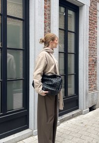 Schwarze geflochtene Leder-Schultertasche mit Reißverschluss, gehalten neben einem hellbeigen Pullover und braunen weit geschnittenen Hosen, vor einer Ziegelmauer.