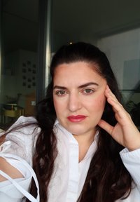 Camisa blanca de cuello con mangas fruncidas, cabello largo y oscuro, maquillaje en tonos neutros y labial rosa sutil. Iluminación suave en el interior.