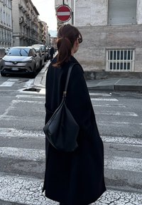 Γυναίκα με μαύρο παλτό και τσάντα στον ώμο, στέκεται σε διάβαση πεζών σε πόλη, κοντά σε σήμα απαγόρευσης εισόδου και παρκαρισμένα αυτοκίνητα.