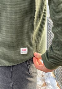 Približnji posnetek osebe, ki nosi zeleno teksturirano Levi's pulover, drži rob puloverja, oblečena v sive kavbojke, v snežnem zunanjem okolju.