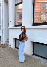 Femme portant des lunettes de soleil orange, un pull noir, un jean bleu clair et un sac à main marron, debout sur un trottoir près d'un bâtiment blanc avec de grandes fenêtres.