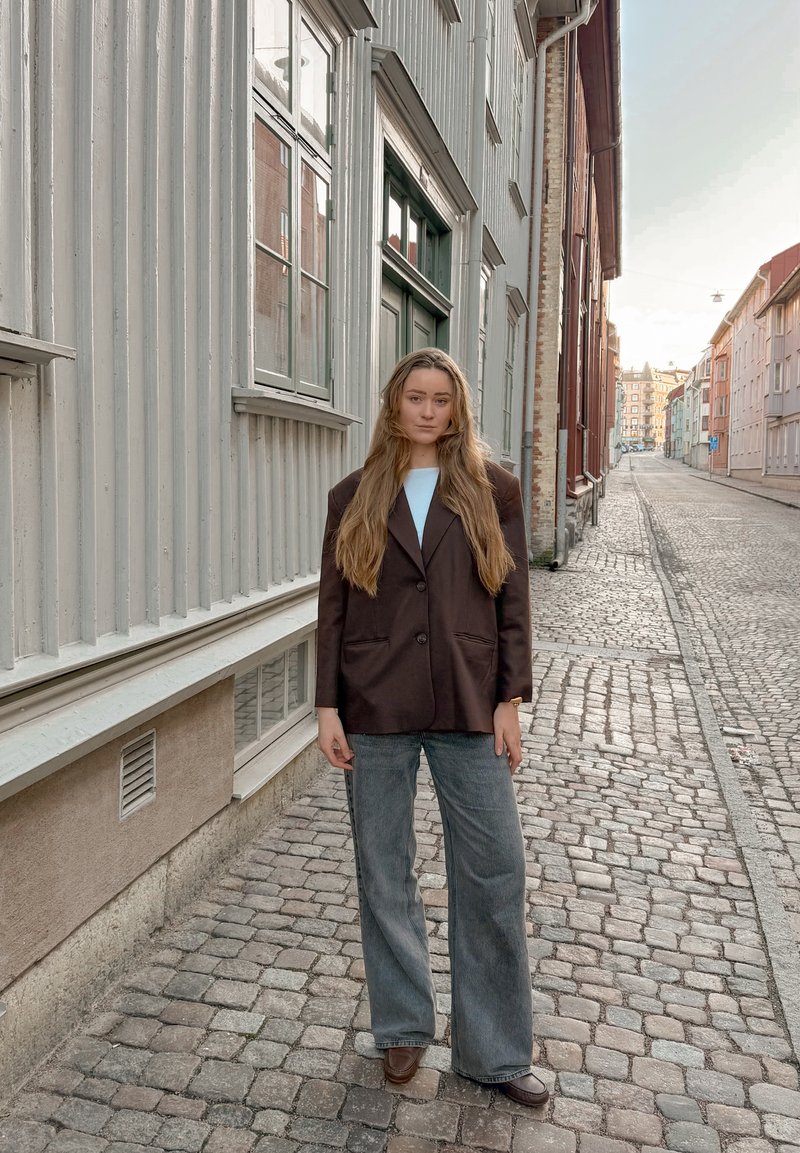 Jeune femme aux longs cheveux portant un blazer sombre et un jean à jambes larges, debout dans une rue pavée bordée de bâtiments de style européen traditionnel.