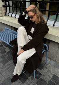 Hnědý oversized kabát, pruhovaná tmavá a bílá košile, krémové kalhoty a tmavé mokasíny. Sluneční brýle z želvoviny. Sedí na modré lavici.