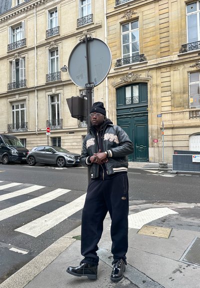 Chaqueta bomber de cuero negra con letras beige, pantalones de chándal negros y botas negras brillantes. Ambiente urbano con edificios y señales de tráfico.