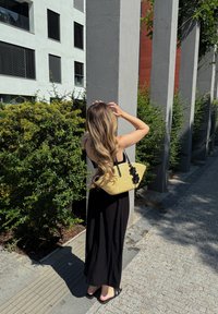 Femme aux longs cheveux ondulés, vêtue d'une robe noire et de sandales, tenant un sac en paille, debout sur un trottoir près de buissons et de colonnes de bâtiment, éclairée par le soleil.