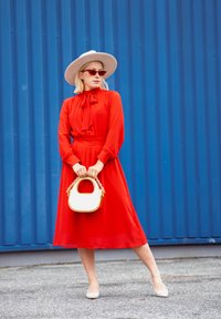 Červené šaty s viazaním pri krku, dlhými rukávmi a voľným strihom. Béžový klobúk a kabelka so zlatým řetězem. Rúžové slnečné okuliare dotvárajú celkový vzhľad.