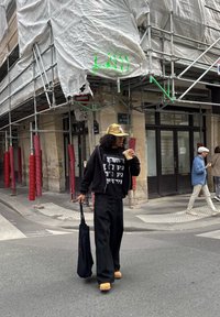 Persona melnā apģērbā un dzeltenā rūtainā cepurē tur kafiju un melnu somu, ejot pa pilsētas ielu ar fonā esošu ēku, kuru aptver skatuves.