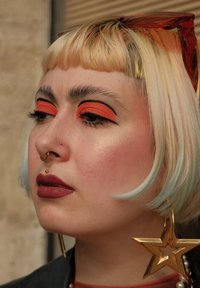 Portret osoby z blond włosami i pomarańczowym cieniem do powiek; posiada złoty kolczyk w kształcie gwiazdy, oversized czerwone okulary przeciwsłoneczne i nagą pomadkę.