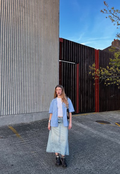 Joven mujer con pelo rubio largo de pie sobre el hormigón cerca de una pared ondulada y paneles de madera verticales oscuros bajo un cielo azul.