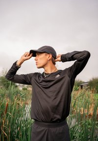 Un giovane che indossa abbigliamento sportivo Adidas nero e un cappellino aggiusta il suo berretto all'aperto vicino a delle alte canne verdi e un cielo nuvoloso.