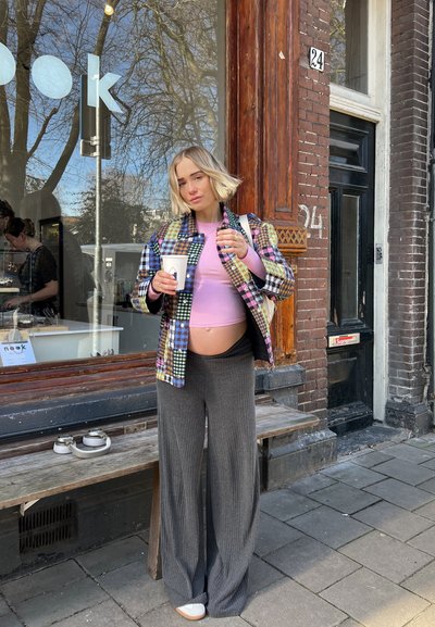 Chaqueta de cuadros colorida sobre una blusa rosa, combinada con pantalones anchos grises. Sosteniendo una taza de café fuera de un café con un banco de madera.