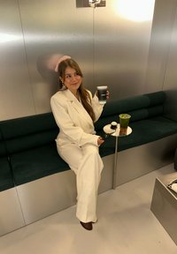 Biały, dopasowany garnitur z fakturowanej tkaniny, siedzący na ciemnozielonej kanapie. Trzyma ciemną filiżankę, obok mały stolik z zielonym napojem. Srebrne tło.