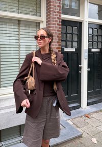 Brązowa, oversized marynarka z szerokimi rękawami, zestawiona z ciemnobrązową bluzką i szarymi szortami. Dodatki to okulary przeciwsłoneczne i mała, beżowa torebka.