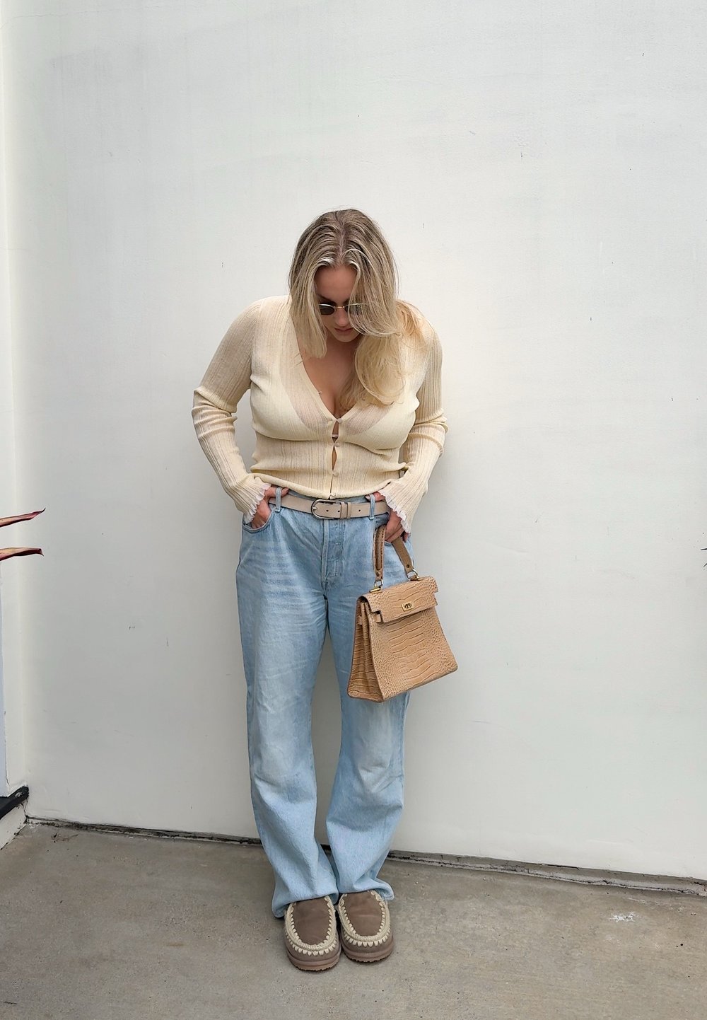 Γυναίκα με μπεζ ζακέτα, ανοιχτό μπλε τζιν και καφέ μοκασίνια, κρατώντας τσάντα ταμπά, στέκεται μπροστά σε λευκό τοίχο κοιτάζοντας προς τα κάτω.