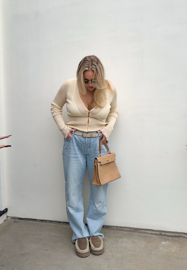 Beige kardigánt, világoskék farmert és barna mokaszin cipőt viselő nő, aki világosbarna kézitáskát tart, fehér fal előtt áll és lefelé néz.