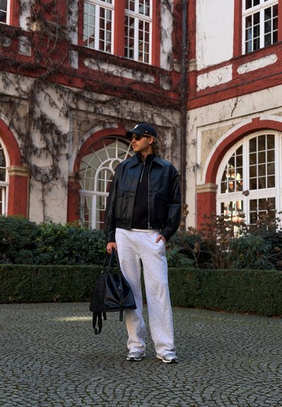 Hombre con chaqueta de cuero negra, gorra negra y gafas de sol, sostiene una bolsa negra, de pie en un patio empedrado frente a un edificio rojo y blanco.