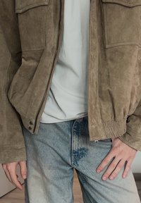 Personne portant une veste zippée en suède beige, un t-shirt blanc et un jean bleu délavé, les mains relâchées le long du corps.