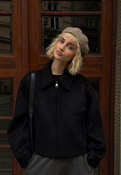 Chaqueta de forro polar negra corta con un gran cuello, cierre de cremallera y bolsillos laterales. Combinada con una gorra de panadero de cuadros beige.