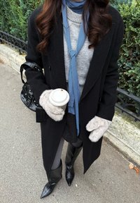 Črn volnen plašč, sivi pulover, modra karo ruta, sive krznene rokavice, črna torbica, črni škornji z visoko peto, drži skodelico kave.