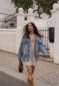 Uma jaqueta de denim azul clara sobre um vestido branco com padrão floral. Bolsa de couro castanho e botas castanhas até ao joelho. Caminho de paralelepípedos.