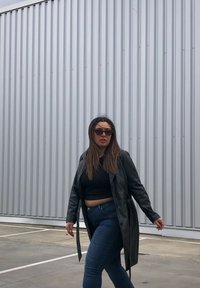 Casaco de trench coat preto em couro sobre um top cropped preto e jeans azuis, com óculos de sol; a caminhar contra um fundo de metal cinza.
