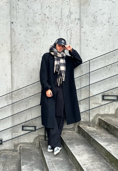 Hombre vestido con un abrigo largo negro, bufanda de cuadros, gorra de béisbol y zapatillas, de pie en unas escaleras de concreto frente a una pared texturizada con un pasamanos de metal.