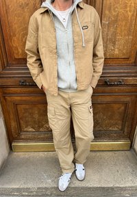 Μπεζ καμβάς μπουφάν πάνω από γκρι φούτερ, συνδυασμένο με μπεζ cargo παντελόνι και λευκά sneakers. Σημαντικά χαρακτηριστικά περιλαμβάνουν φερμουάρ και λογότυπα.