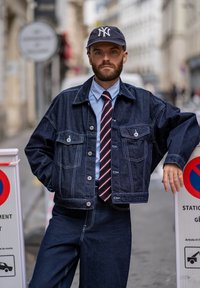 Traper jakna s prednjim džepovima, plava košulja, prugasta kravata i mornarska kapa. Model se oslanja na znak zabranjenog parkiranja. Pozadina ulične scene.