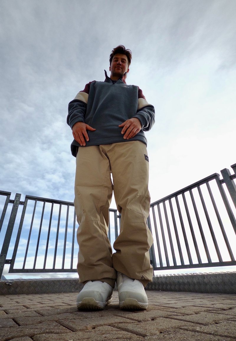 Un hombre con una sudadera y pantalones holgados está de pie en una plataforma pavimentada, detrás de una barandilla metálica, visto desde abajo y con un cielo nublado de fondo.