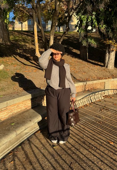 Una mujer con un suéter gris y una bufanda marrón está de pie al aire libre, sosteniendo un bolso de tejido oscuro, con pantalones de pierna ancha marrones y zapatos grises.