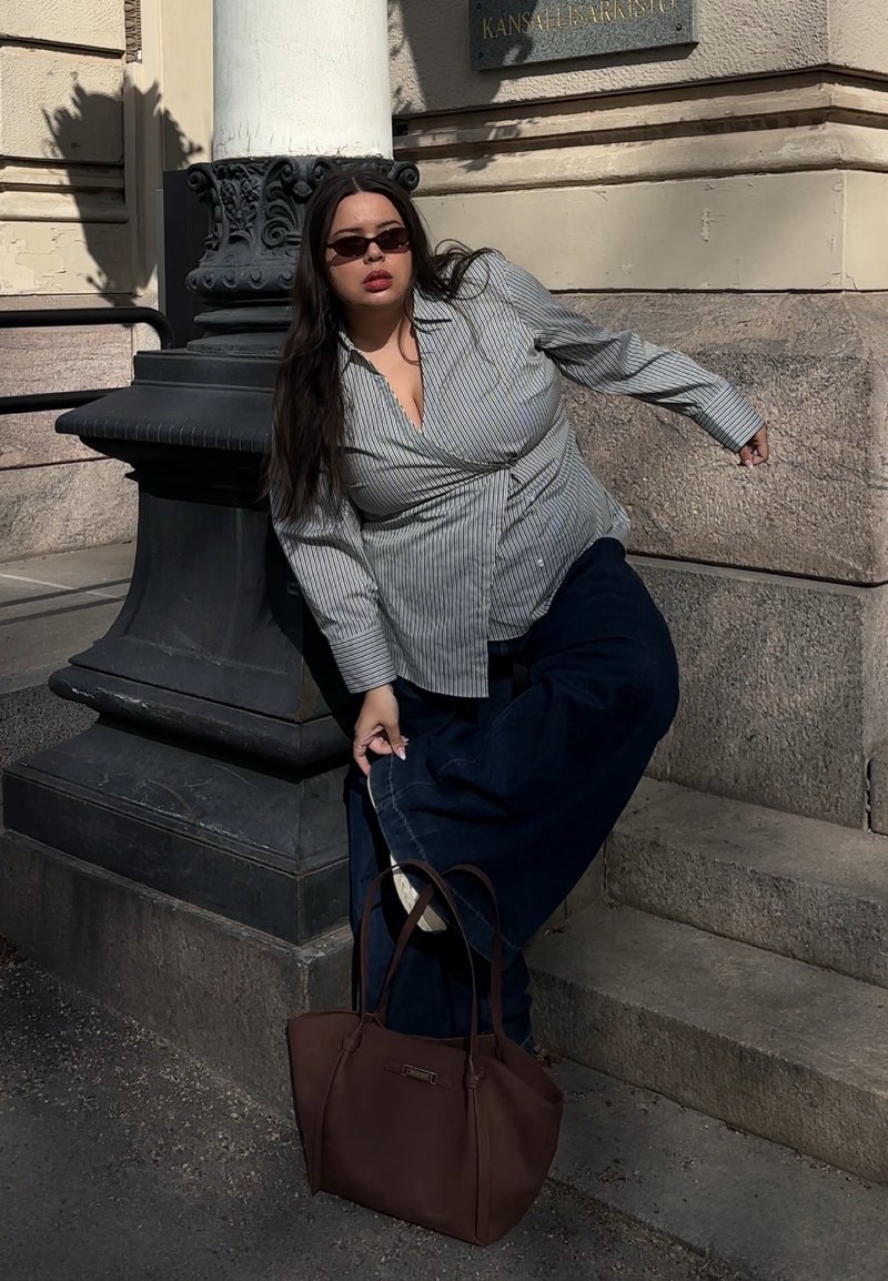 Femme portant des lunettes de soleil, une chemise rayée et un pantalon sombre, ajustant sa chaussure tout en s'appuyant contre un pilier en pierre à l'extérieur.