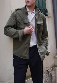 Olivgrüne Militärjacke im Stil mit Knopfleiste, Schulterklappen, zwei Brusttaschen und lässiger Passform über einem weißen Hemd und dunklen Hosen.