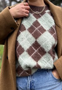Pull à motif losanges marron et vert menthe en matière pelucheuse, porté sous un manteau marron, associé à un jean en denim.