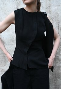 Top senza maniche nero in tessuto texture, con colletto alla mandarina e orlo asimmetrico. Indossato sopra pantaloni neri.