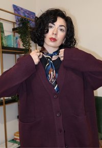 Kaštanový kardigan s gombíkmi na prednej strane a vreckami, doplnený vzorovaným modro-hnedým šálom zviazaným okolo krku, v interiérovom prostredí.