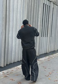 Chándal deportivo negro con ribete blanco, que incluye una chaqueta con cremallera y pantalones holgados. El sujeto lleva auriculares intrauditivos.