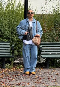 Jeansjacke, schwarze Weste, weißes Hemd, lockere blaue Jeans, beige Clogs, Sonnenbrille und eine braune gemusterte Tasche. Grüne Blätter im Hintergrund.