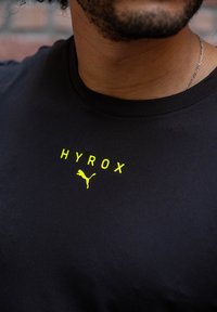 Must t-särk, millel on erksate kollaste tähtedega "HYROX" ja väike kollane Puma logo. Sujuv kangas ja ümara kaelusega disain.