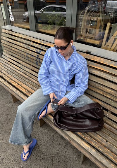Camisa de botones a rayas azul y blanca, jeans de mezclilla claros, chanclas azules, bolsa de cuero marrón al lado, sentado en un banco de madera.