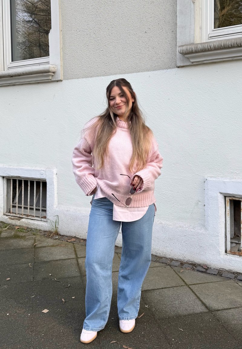 Jeune femme en pull rose, jean bleu clair et chaussures blanches se tient sur le trottoir en tenant des lunettes de soleil près d'un bâtiment blanc aux fenêtres grillagées.