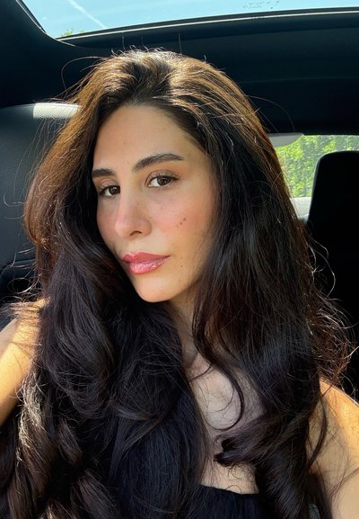Acercamiento de una persona con cabello largo y ondulado negro, piel suave y un maquillaje natural. Se puede ver el interior de un coche negro de fondo.