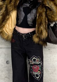 Άτομο που φοράει ένα μαύρο κοντό τοπ με φτερά από στρας, παλτό από γούνα, ζώνη με καρφιά και μαύρο παντελόνι με μεγάλο κόκκινο τρι αντάφυλλο και μπαλωμένο σταυρό.