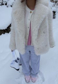 Manteau en fausse fourrure blanc, cardigan rose clair avec boutons, pantalon de survêtement gris et chaussures rose clair. Tient des patins à glace blancs avec des accents noirs.