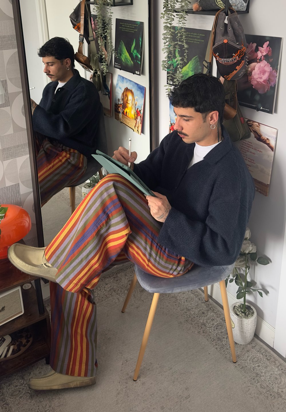 A person dressed in a dark knitted sweater and multi-coloured striped wide-leg trousers, seated on a grey chair, creating a drawing on a light green tablet.